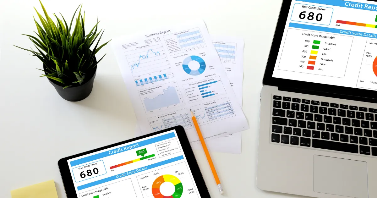 A laptop, tablet, and business reports displaying credit scores and financial charts on a desk.