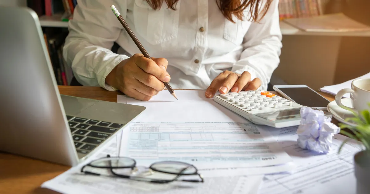 A person writing notes with a pencil while using a calculator and laptop at a desk.