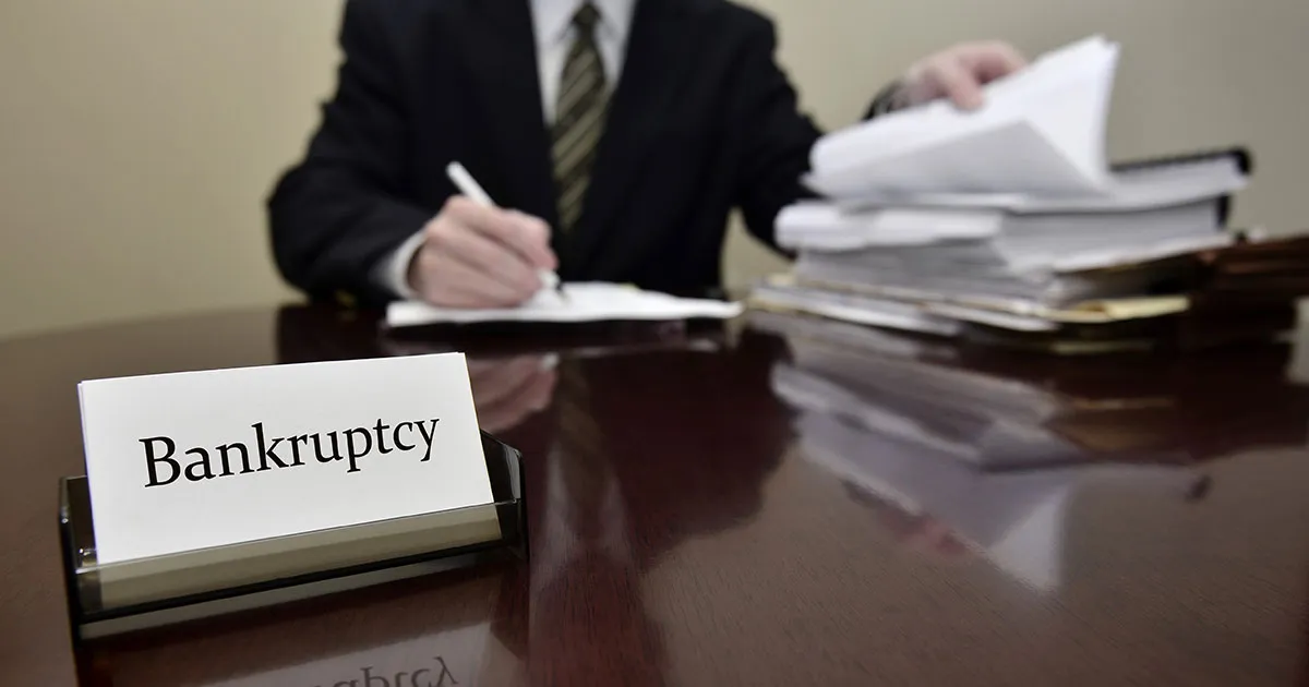 A sign that reads "Bankruptcy" on a desk with a lawyer reviewing documents in the background.