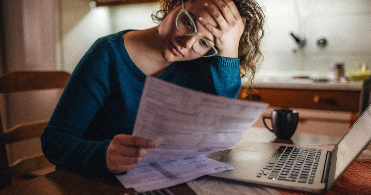A stressed woman holding her head while reviewing bills at a table with a laptop.