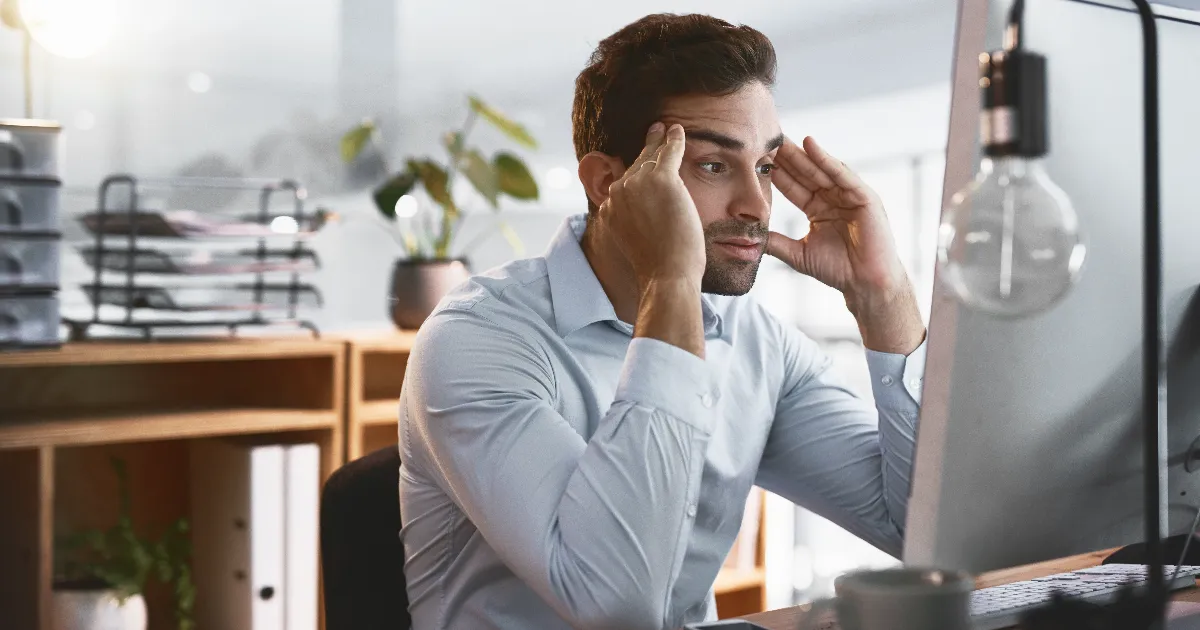A stressed man rubbing his temples while looking at a computer screen