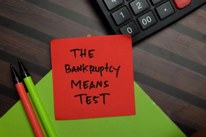 A red sticky note with the handwritten text "THE BANKRUPTCY MEANS TEST" next to a calculator and pens.