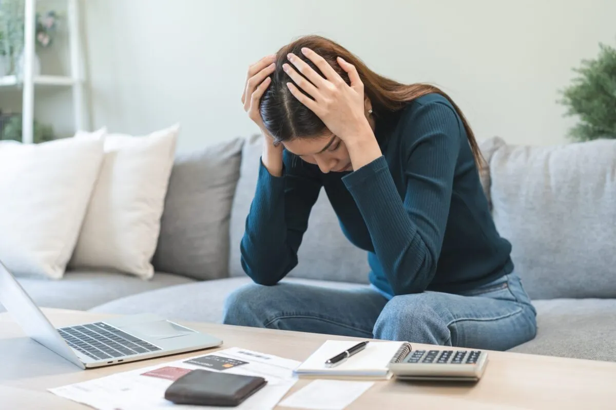 A stressed woman holding her head while sitting on a couch with bills and a laptop on the table.
