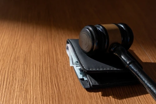 A wooden judge's gavel rests on top of a black leather wallet filled with cash on a wooden table.